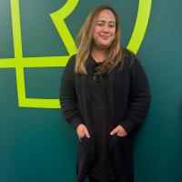 Marie stands against a dark green wall with the RDP logo. She has long dark blonde hair and wears a long back sweater.