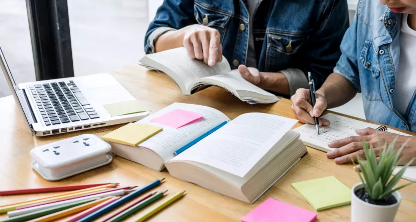 Two students study at a desk covered in books, notepads, and pencils. Their faces are not seen in the photo.