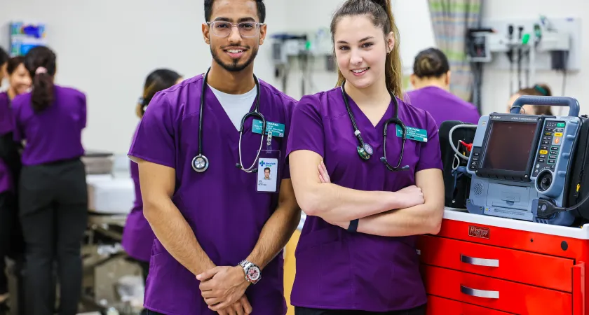 Two students in purple scrubs standing confidently beside each other in a lab smiling at the camera.