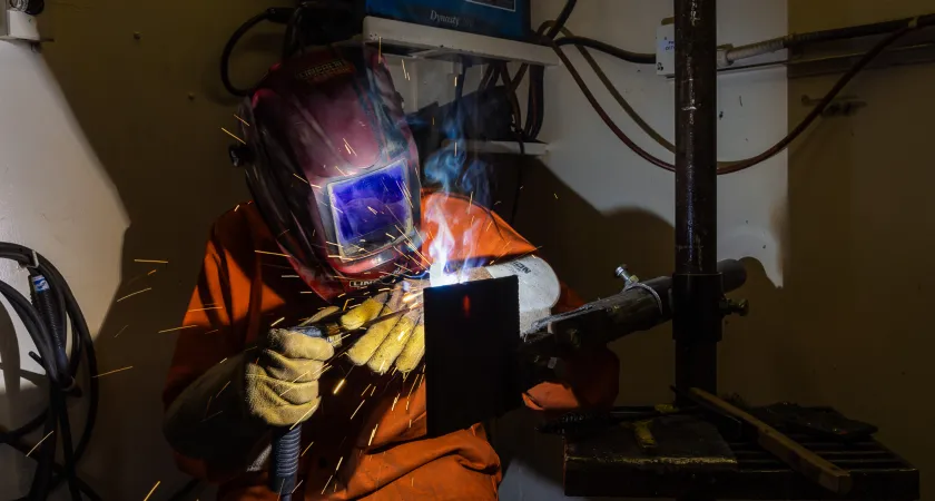 A welder in protective helmet and gloves, welds a piece of metal.