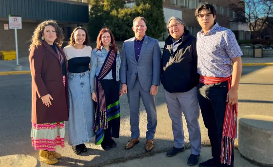 A group of students, admin, and community members stand together. Two wear the Métis Sash and two wear Ribbon Skirts. 
