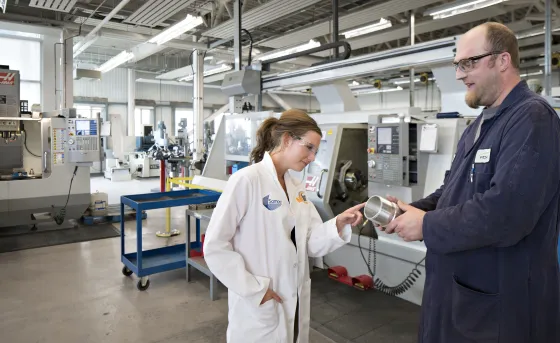 A female student in a lab coat examines a part being shown to her by a male lab tech in the CIM-TAC
