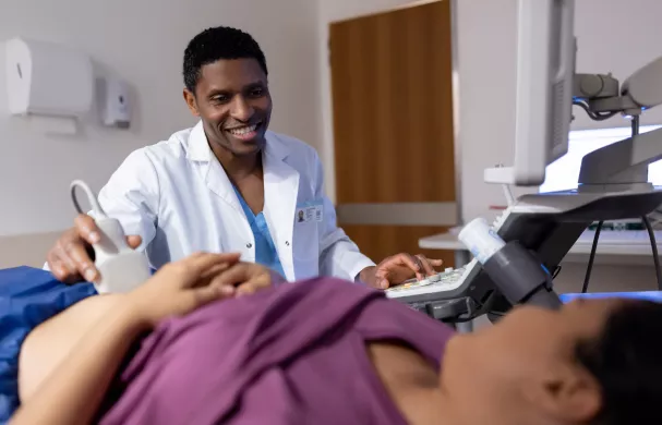 A sonographer smiles while using an ultrasound wand on a patient abdomen.