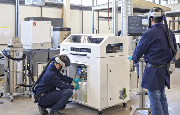 Two men wearing blue lab coats and protective helmets are working with the ExOne 3D Printer