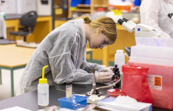 A female student in protective gear works with small bottles of chemical compounds in a lab