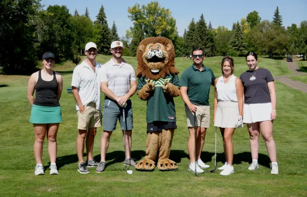 A group of golfers, student athletes, and Rufus the lion stand on a golf course fairway