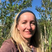 Elissa smiles slightly on a bright day surrounded by green shrubs and grasses. She wears a headband, hoop earrings, and a coruroy jacket.