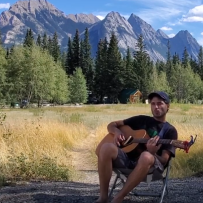 Etienne LeBel sits in a lawn chair playing a guitar. There is a beautiful mountainscape behind him. 