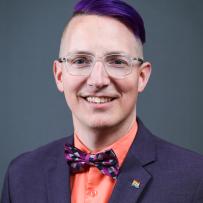 Robbie has purple hair swept to one side, glasses, and wears a purple suit, bright orange shirt, and purple bowtie.