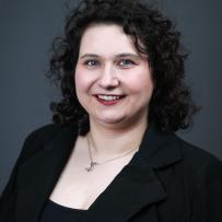 Jacq Patterson has dark curly hair, wears a black outfit and smiles at the camera while posing against a dark grey background