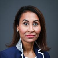Melina wears a navy blue blazer with white accents and a white shirt. She poses against a dark grey background and looks into the camera