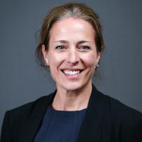 Tristin Brisbois wears a black blazer and blue shirt underneath. She poses against a dark grey background and smiles at the camera