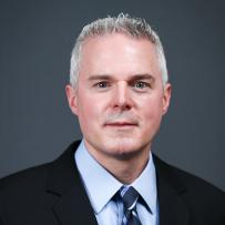 Dustin Quirk wears a dark suit, light blue shirt, and dark blue tie. He poses against a dark grey background and looks into the camera
