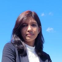 Taslima looks slightly down towards the camera with a neutral face. The clear blue sky behind her