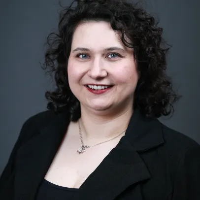 Jacq Patterson has dark curly hair, wears a black outfit and smiles at the camera while posing against a dark grey background