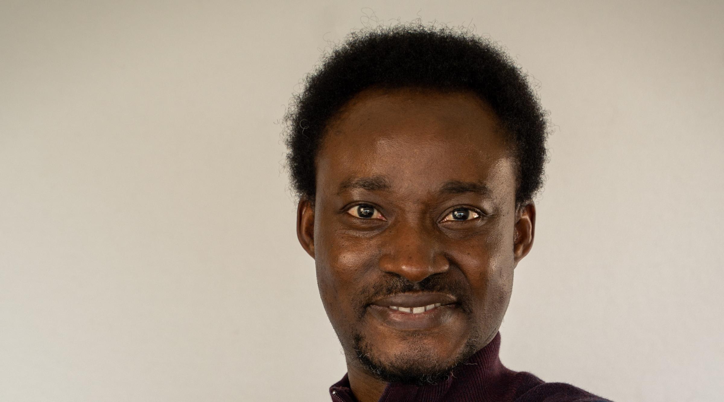 Fola stands with arms crossed, smiling at the camera. He wears a black shirt.