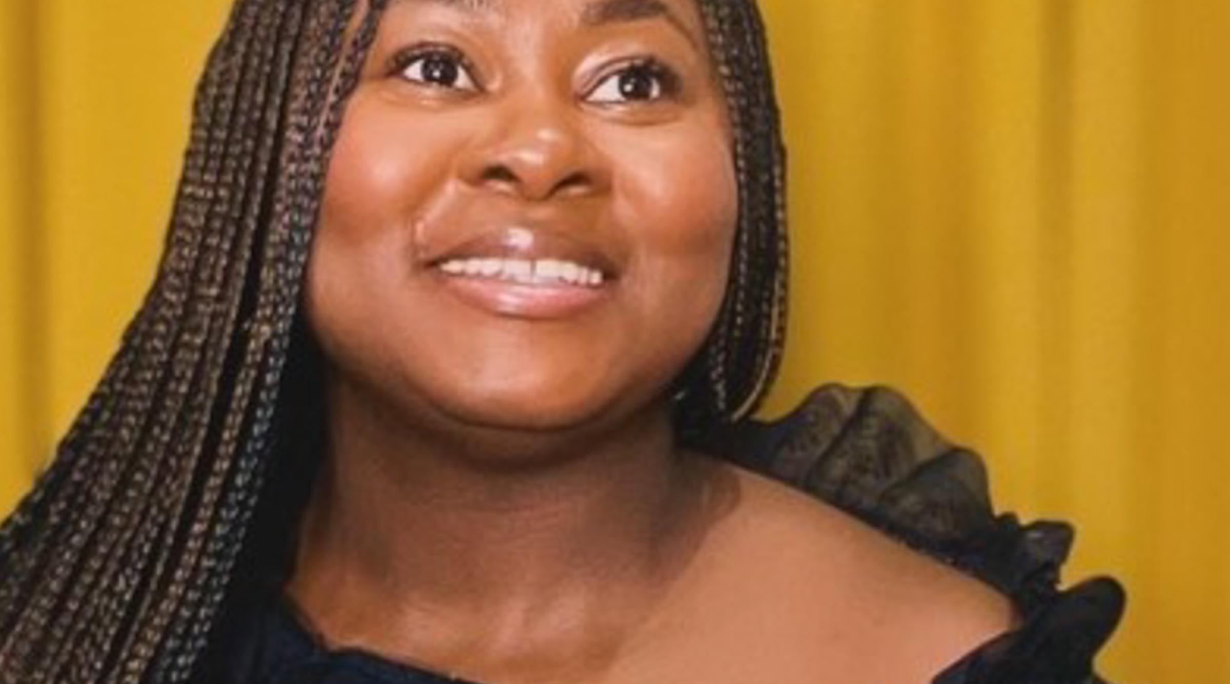 Oluwaseyifunmi smiles looking upwards. Her hair falls in long braids over her black dress.