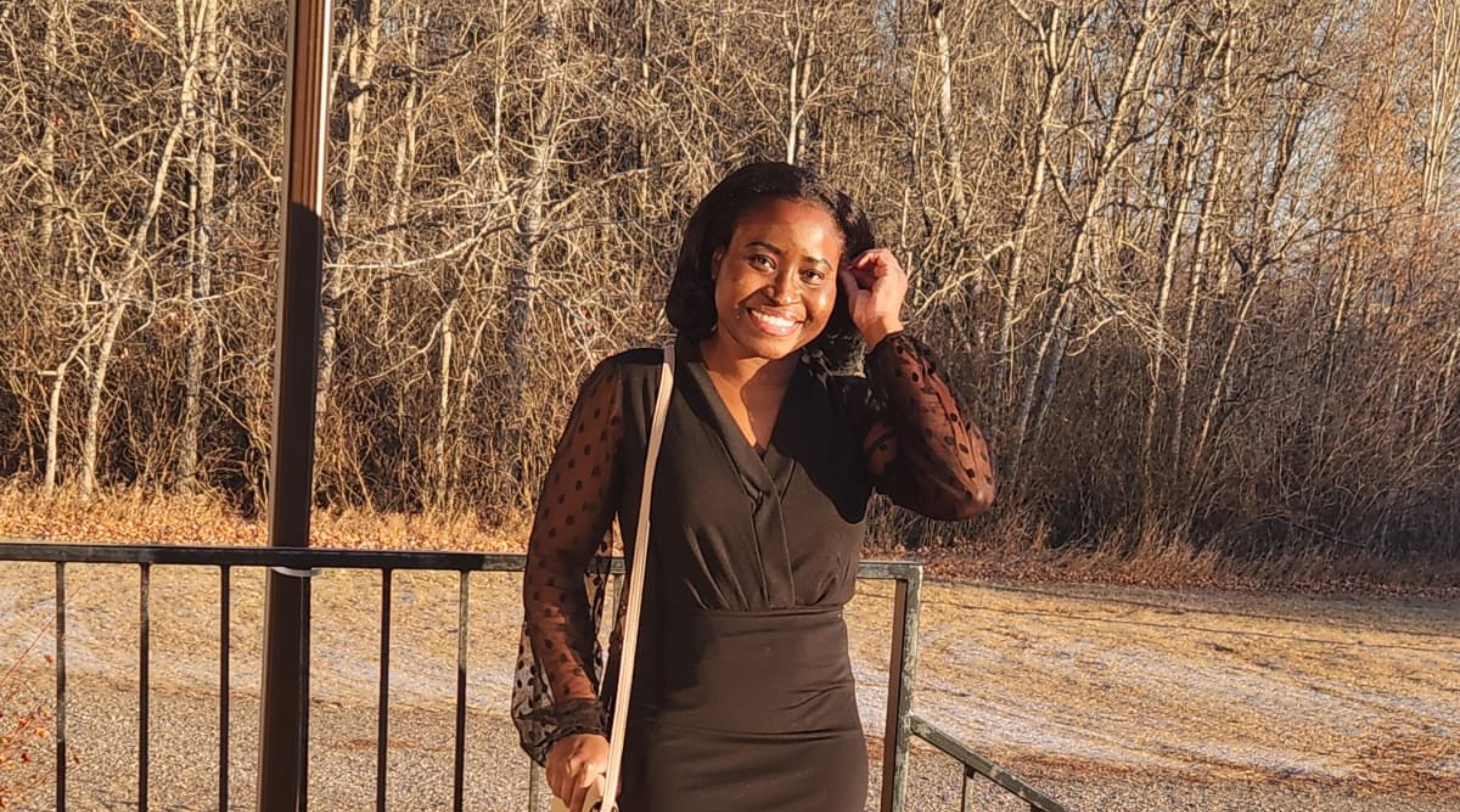 Rys-Ann wears a black dress with a grey purse on her shoulder. She smiles while running her fingers through her hair. She is standing on concrete steps outside in bright late-day sunshine.