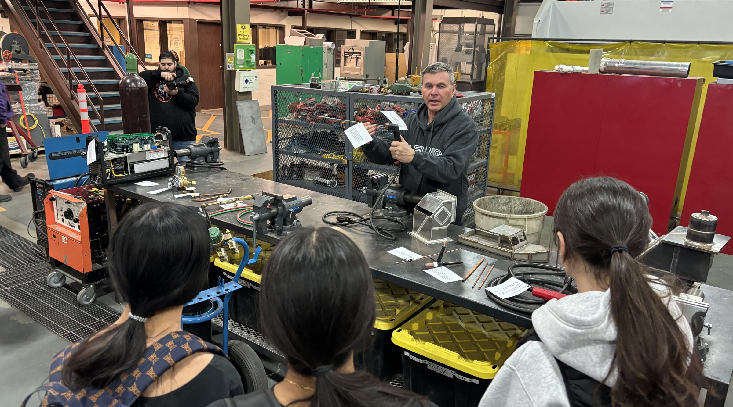 A male instructor stands behind a desk full of tools explaining how they work to a group of three female students