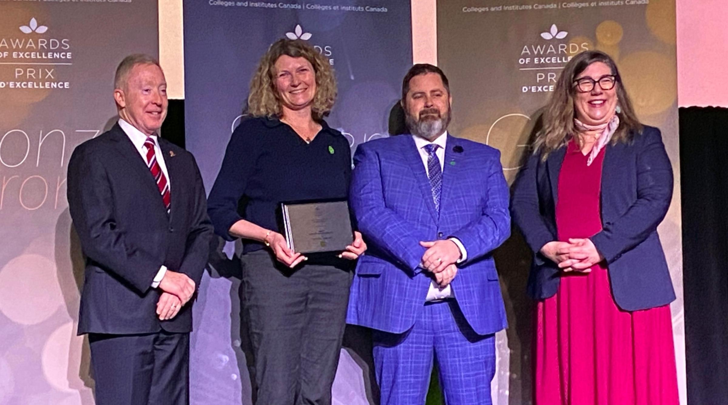 A smiling group of stands around Alicia Caffereta who holds her recent award