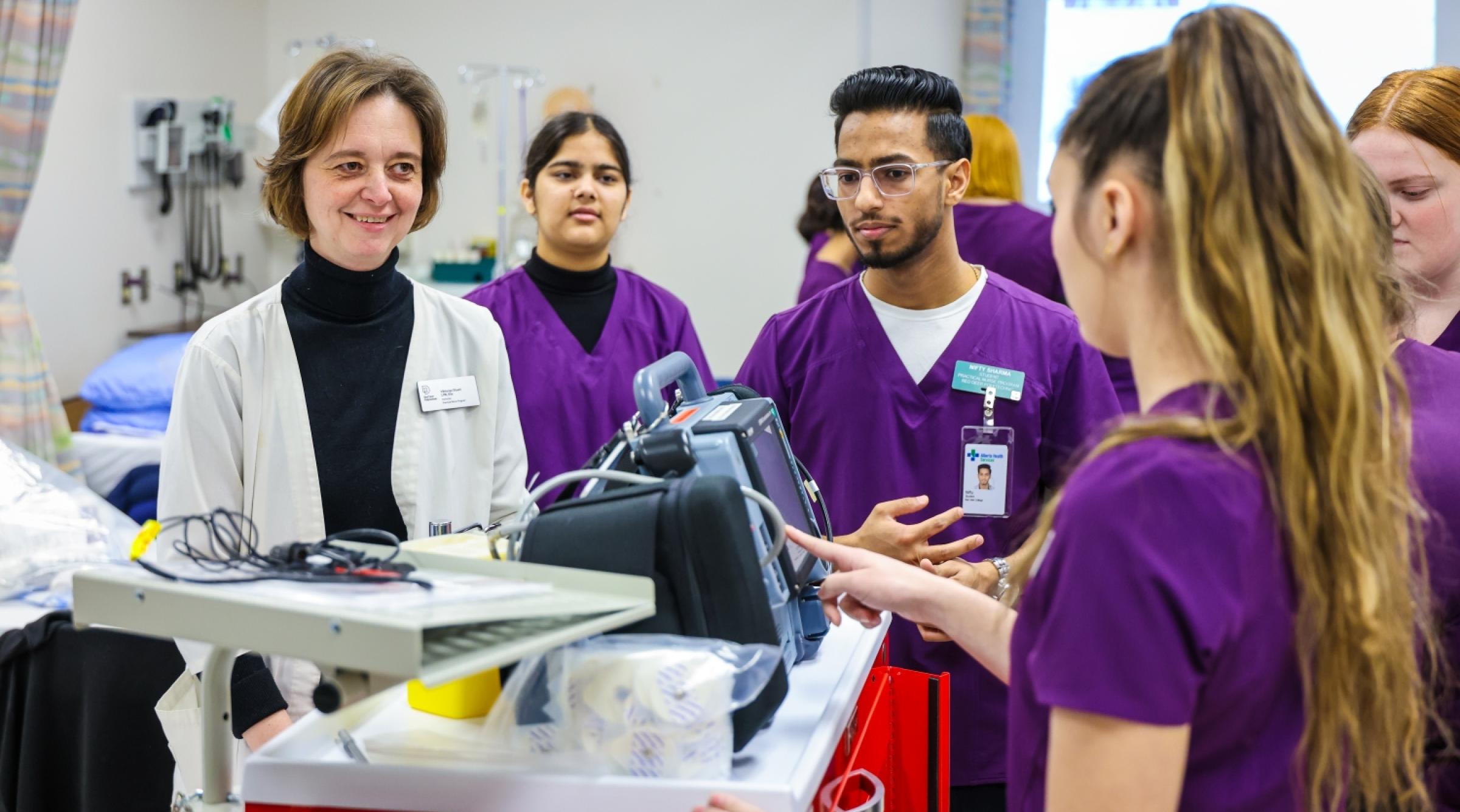 A group of nursing students in purple scrubs working on equipment while instructor watches