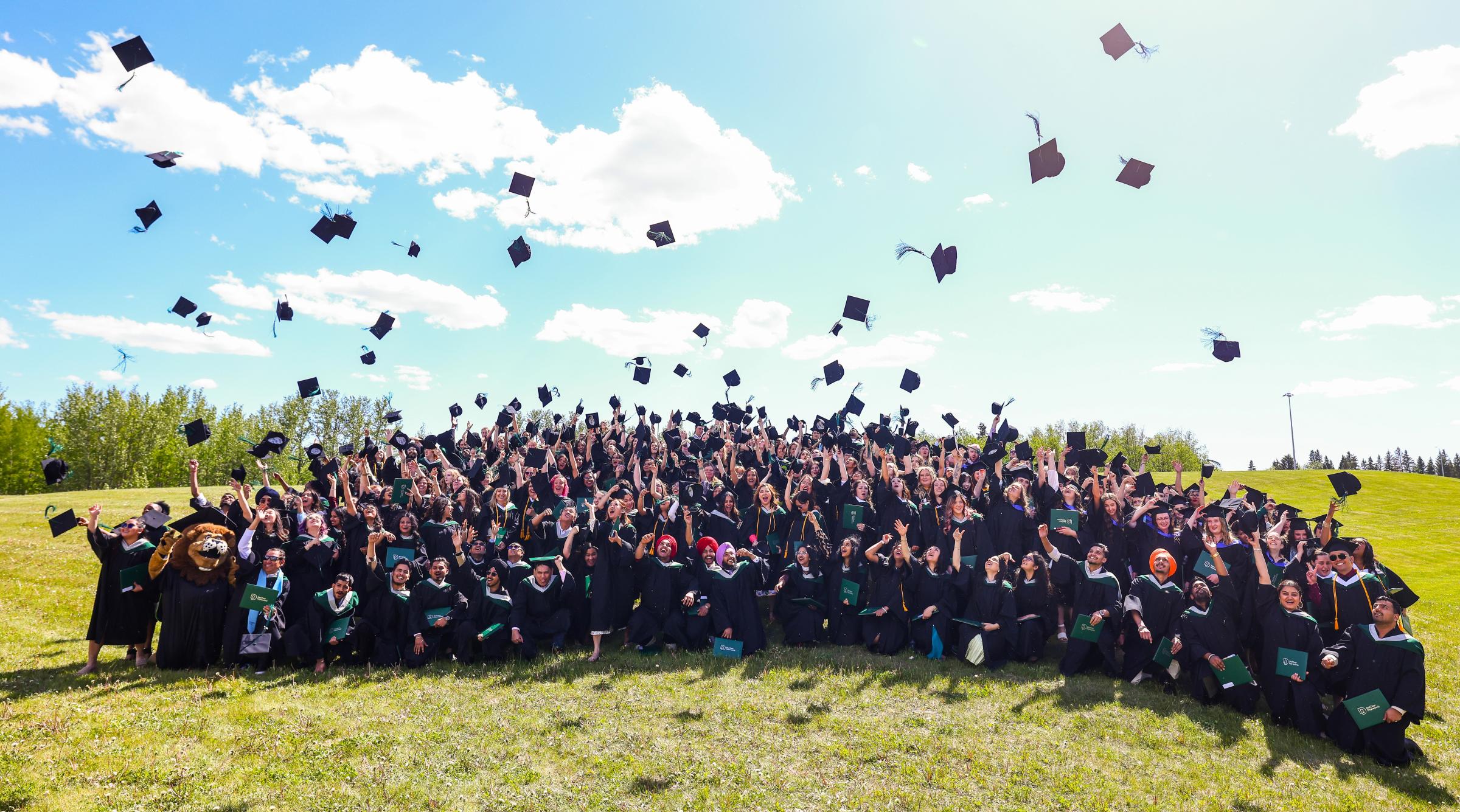 A large group of students in graduation gowns are throwing their grad caps into a clear blue sky above them
