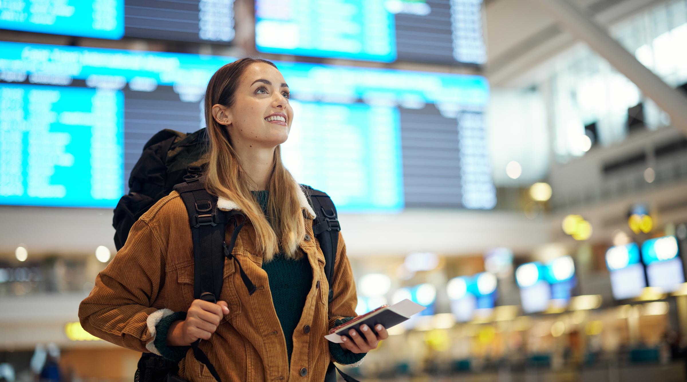 A student looks up at airport status boards with an excited smile. Thye have a large backpack on and are holding their passport.