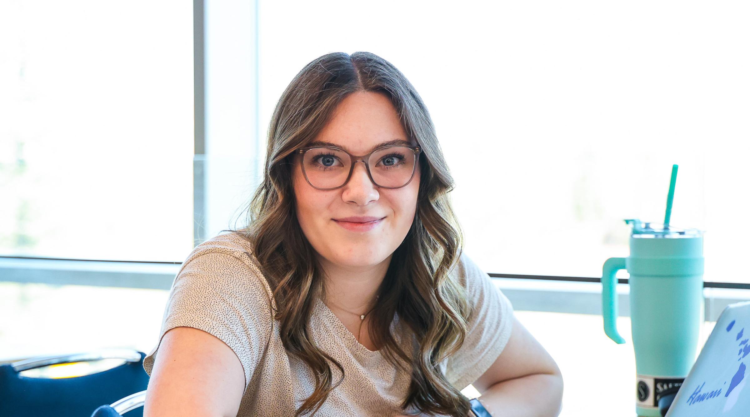 A student with long brown hair sits in front of brightly lit windows at a table. Their books, laptop, and large water jug sit on the table in front of them.
