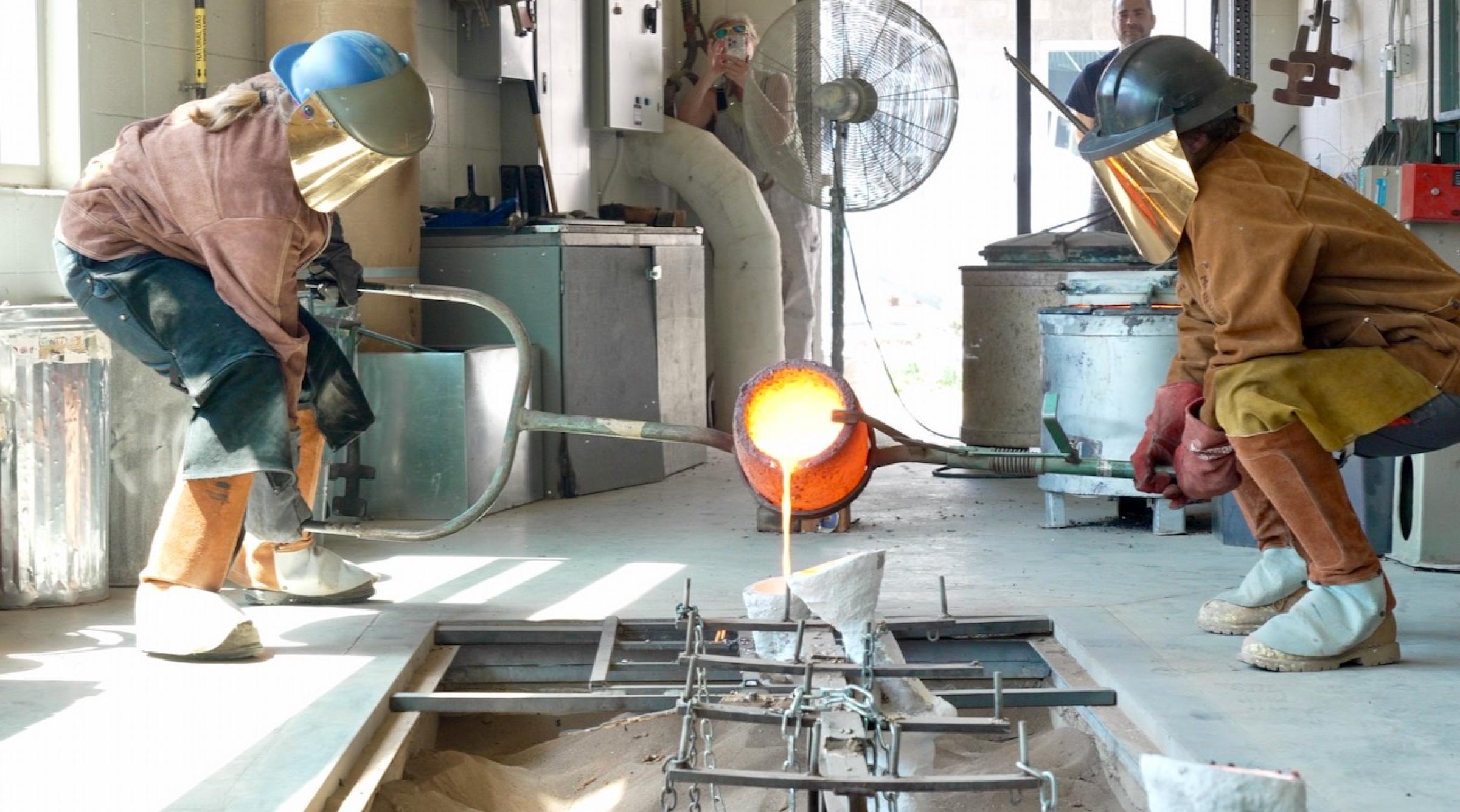 Two people in protective suits pour molten bronze into a mold in a workshop