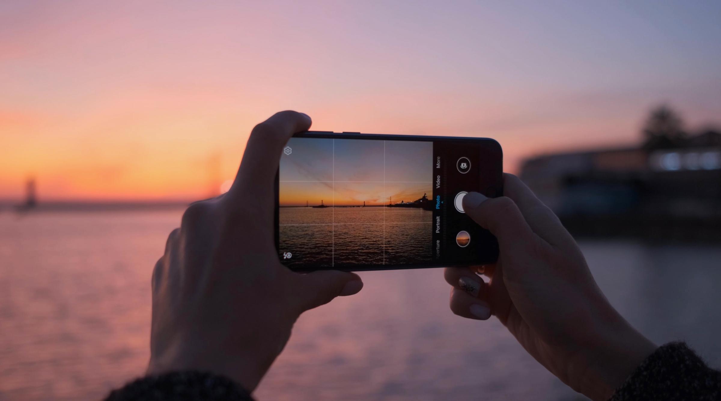 A view through a phone camera held by two hands, taking a picture of an ocean at sunset.