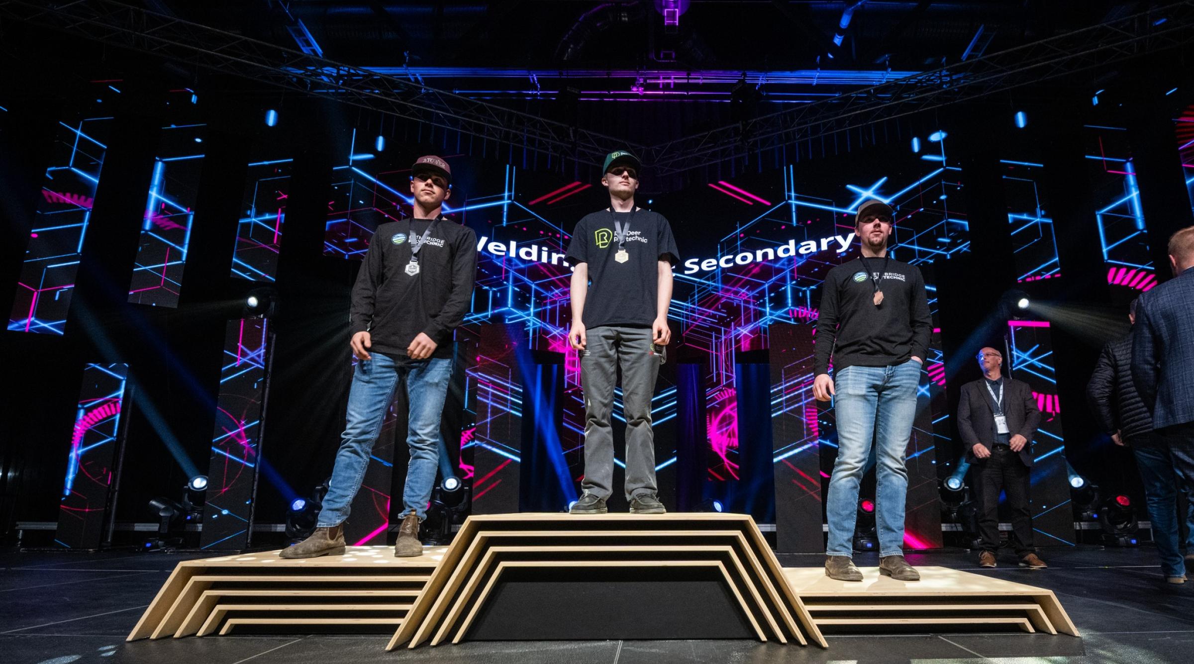 Lucas Stang (RDP Welding student) stands with his gold medal on a podium with two other welders at Skills Canada Alberta 2025
