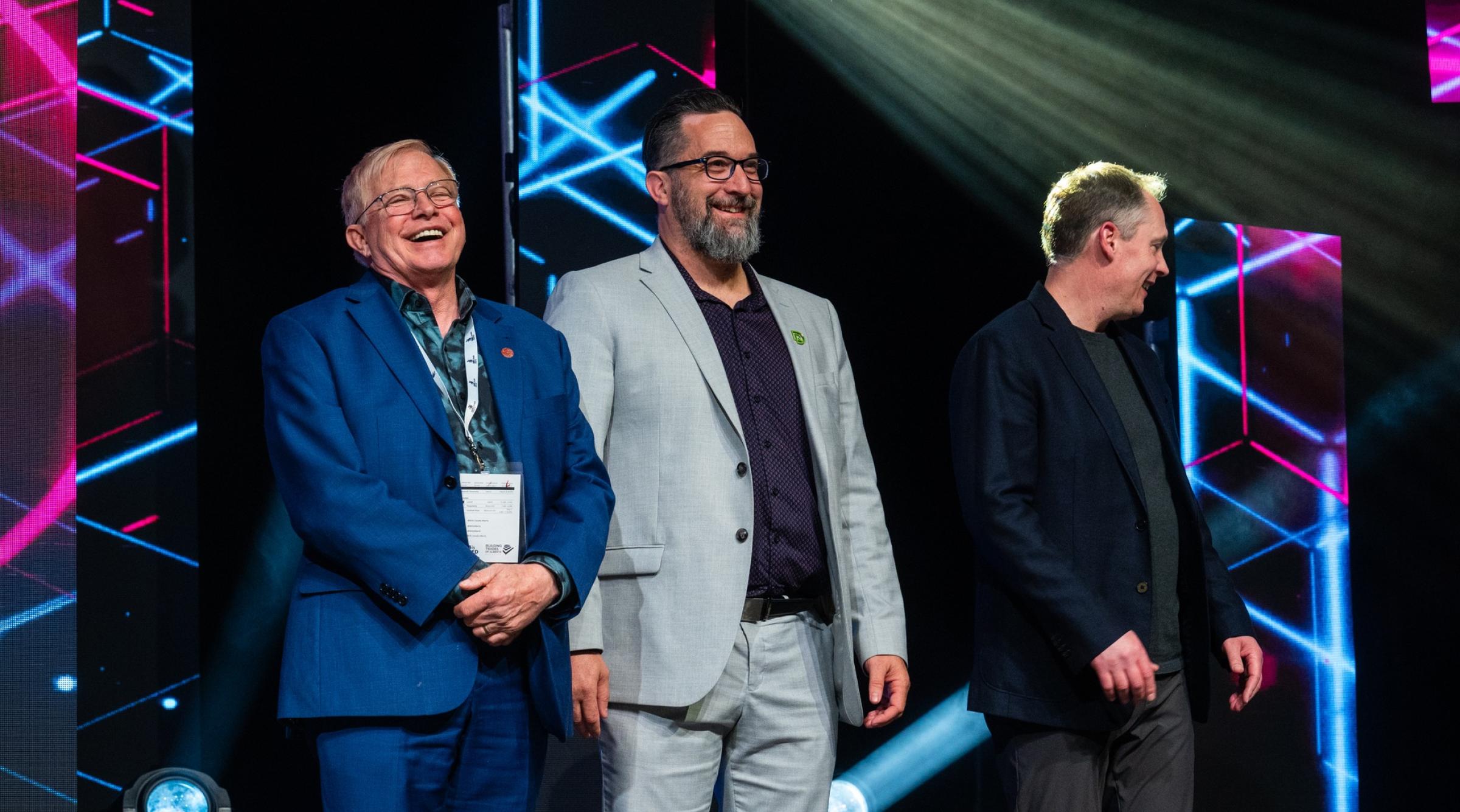 David Pye in a grey suit and black shirt stands smiling on stage with two other suited men at Skills Canada Alberta 2025