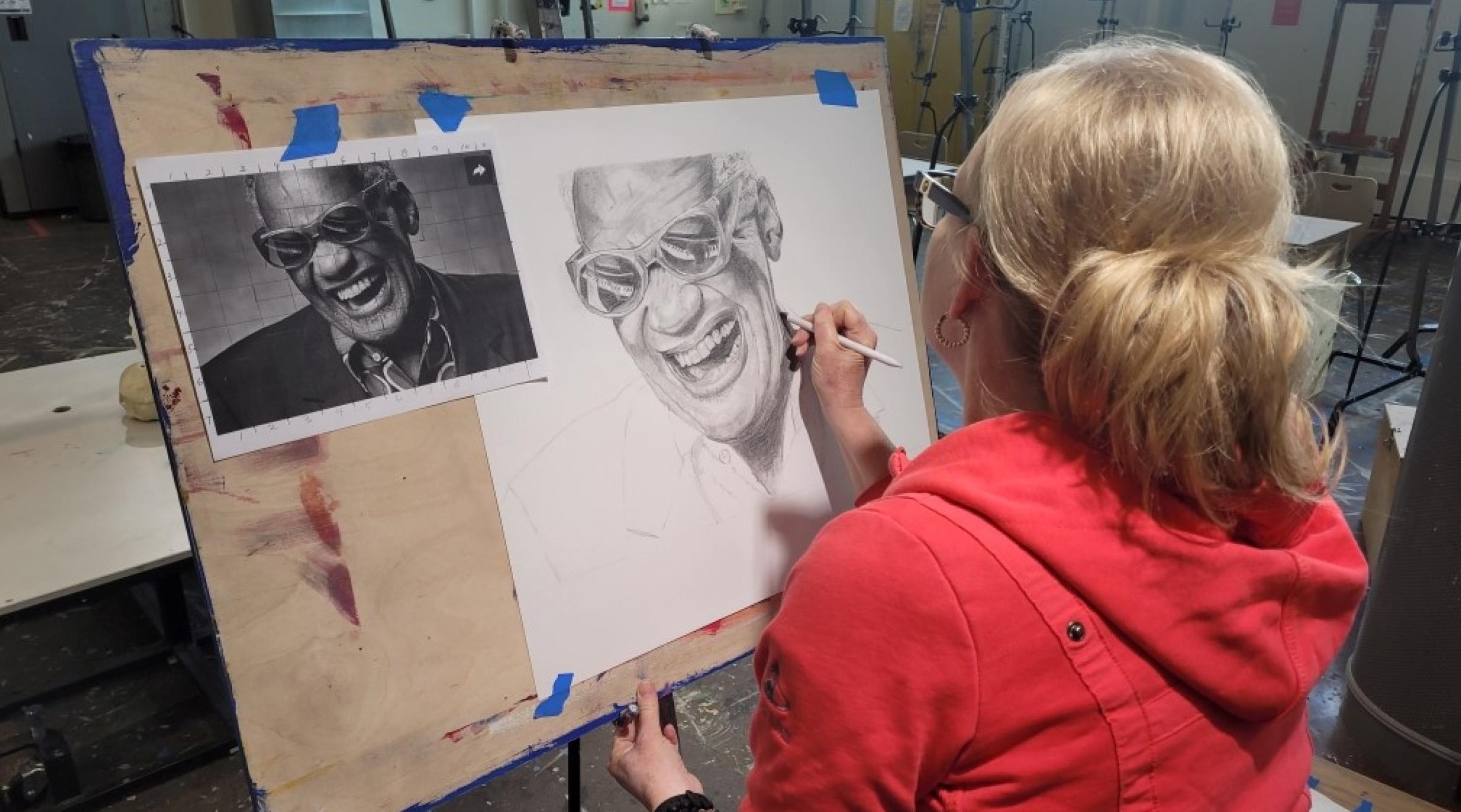 A woman in a red hoodie sits in an art studio. She is sketching a picture of Ray Charles using a pencil