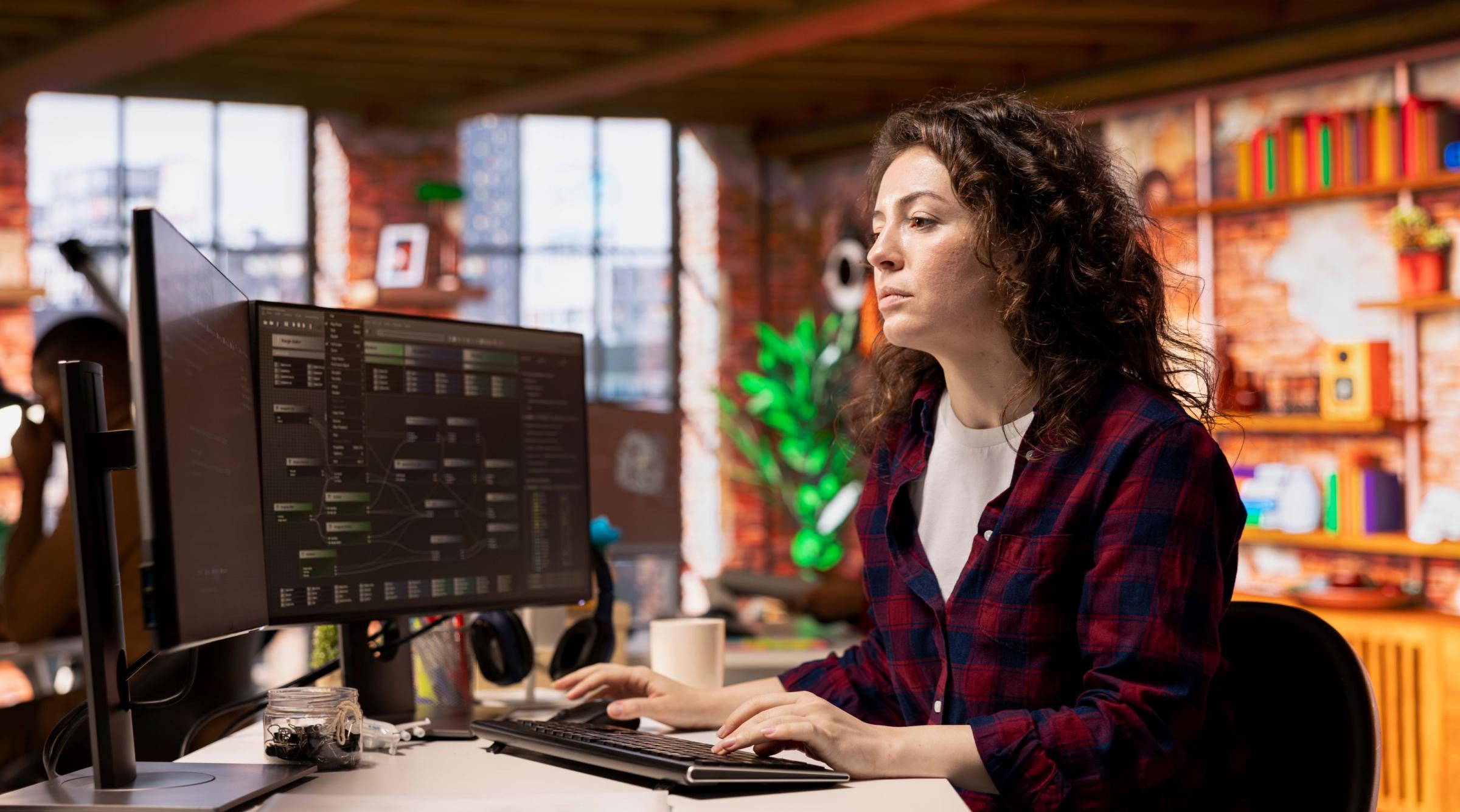 A woman with long brown hair sits in an office in front of a computer with two monitors