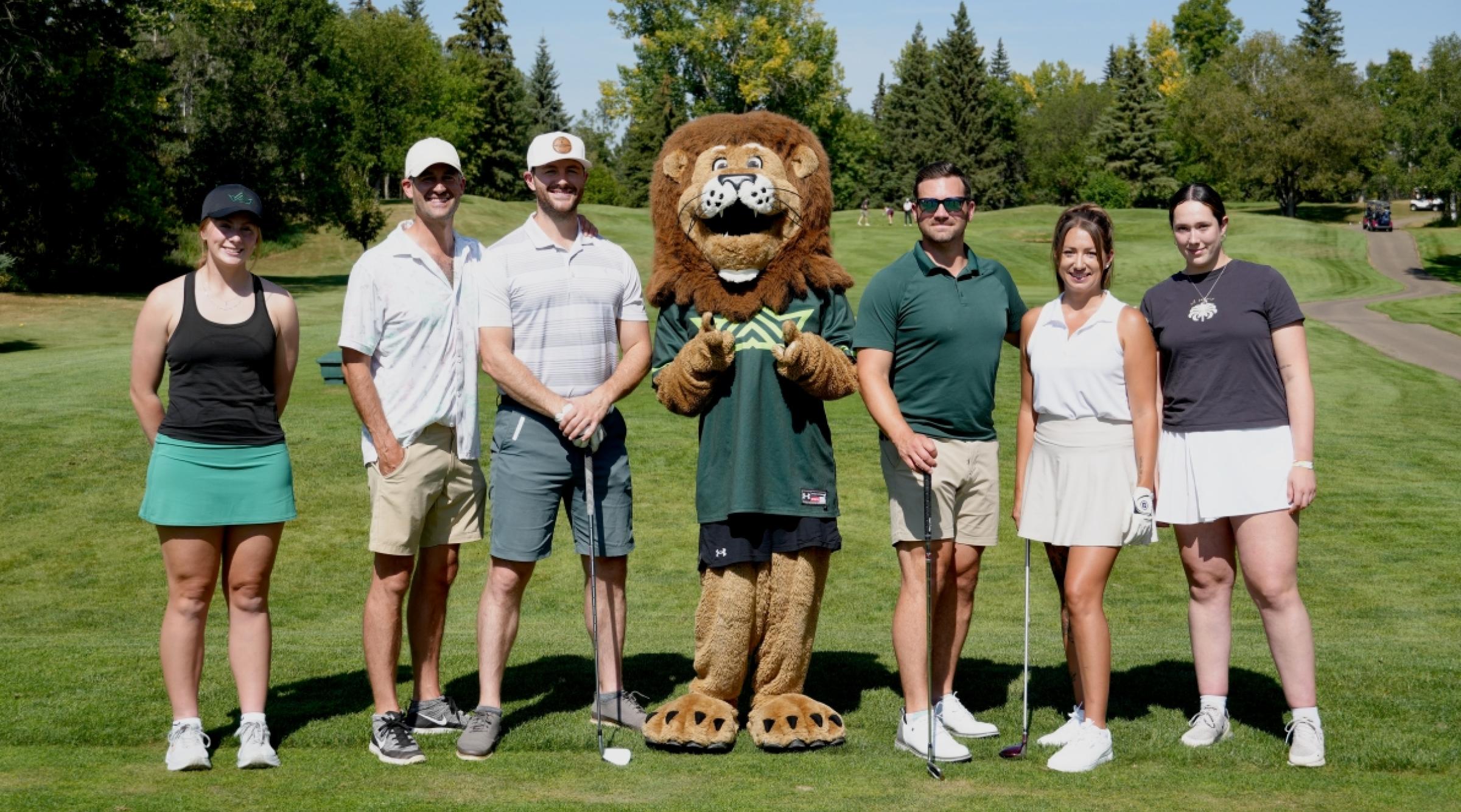 A group of golfers, student athletes, and Rufus the lion stand on a golf course fairway