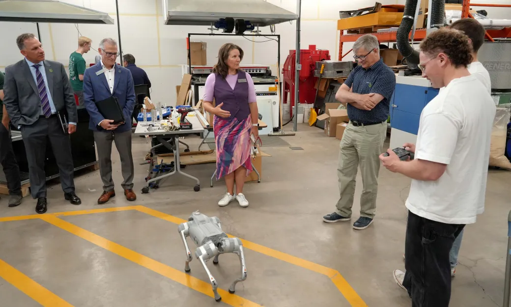 A group stands around a robot dog, Dr. Woof in a workshop. One student holds the controller for Dr. Woof