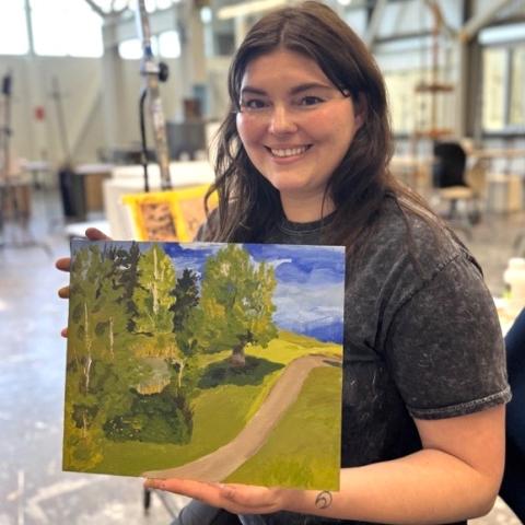 Alex King smiles holding a landscape painting she's made during the Series Summer Art workshops