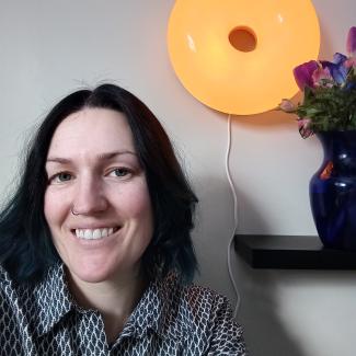 Taylor, with long black hair, smiles in front of a donut shaped light and a blue vase with purple and pink flowers.