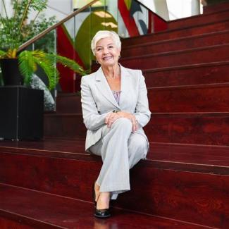 A smiling Gail Surkan, sits wearing a grey pantsuit on some dark wooden steps