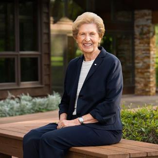 Joan Donald smiles while sitting on a wooden bench in a dark blue pant suit in front of a house