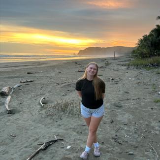 Jessie horning has long blonde hair and smiles on a beach in front of the ocean and the setting sun