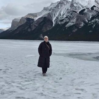 Lisa wears a black jacket and white ballcap standing in the snow in front of a grand mountain