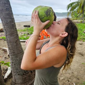 Jennine Baughman drinks a coconut on the beach with palm trees and water behind her