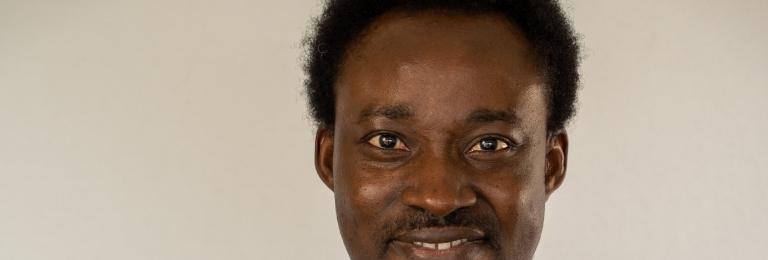 Fola stands with arms crossed, smiling at the camera. He wears a black shirt.