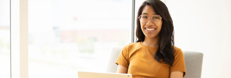 Smiling student sitting in a chair with a laptop on her lap in front of a bright window
