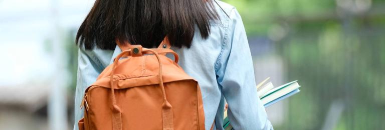 A student with long black hair wearing a long sleeve blue shirt and a brown backpack faces away from the camera looking at green trees in the background.