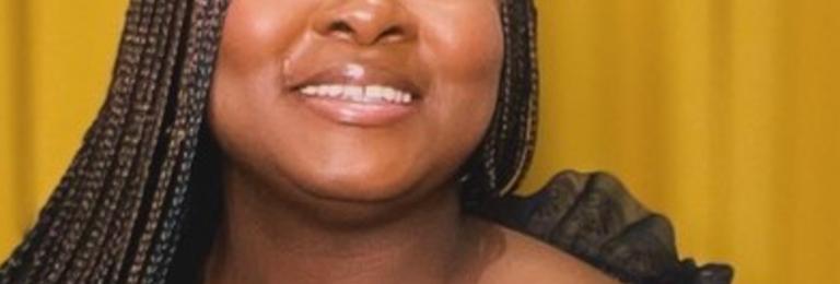 Oluwaseyifunmi smiles looking upwards. Her hair falls in long braids over her black dress.
