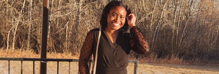 Rys-Ann wears a black dress with a grey purse on her shoulder. She smiles while running her fingers through her hair. She is standing on concrete steps outside in bright late-day sunshine.