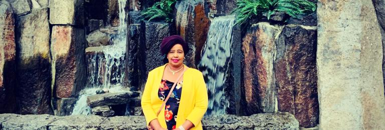 Zanaib stands in front of a large rock formation, wearing a flowered dress and yellow jacket.