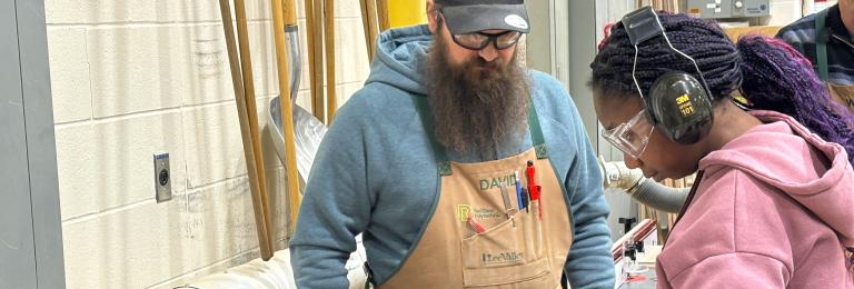 A bearded faculty member watches as a young female student cuts some wood on a large table saw.